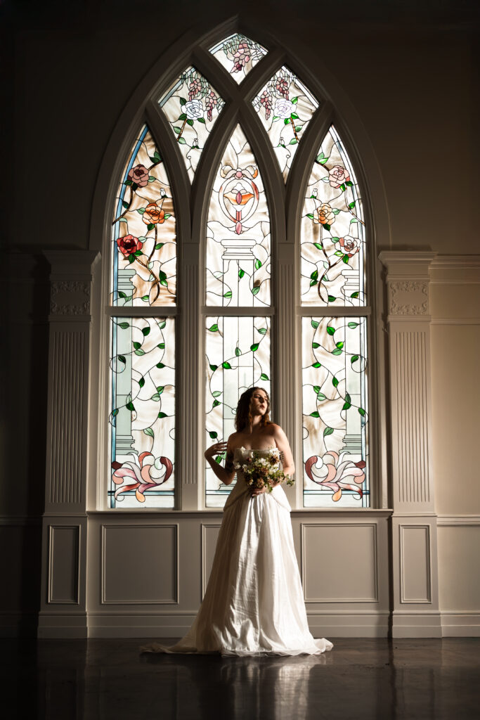 Wide-angle image: bride in gown framed by classic architecture of a large floral stained glass window in Northampton House, blending old-world charm with bold editorial style.