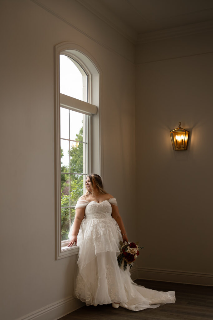 "Fine art Utah bridal session inside The Magnolia Event Center, bride in lace gown from Bridal Brilliance SLC holding bouquet by the window."