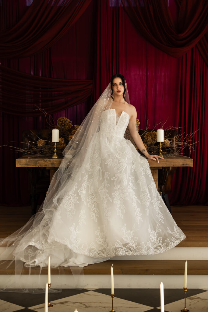 grand ballgown set against a wooden table with dried hydrangeas and twigs that have been arranged as a showstopping centerpiece. Maroon drapes cascade in the background