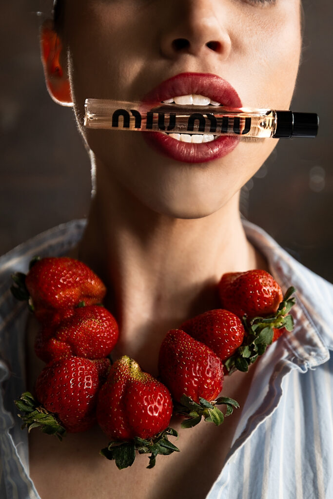 Miu Miu Miutine Eau de Parfum clenched between the teeth of a model with a vibrant necklace of strawberries around her neck, bold lipstick, and light blue and white striped collared shirt. An Editorial campaign picture.